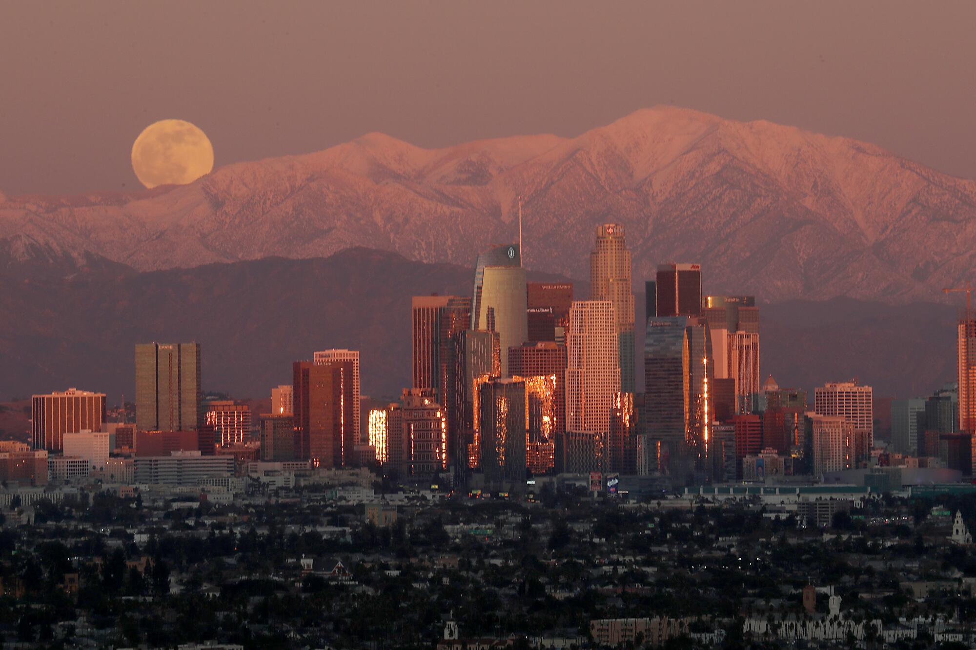 A full moon rises over the snow-capped San Gabriel Mountains and the skyline of downtown Los Angeles.