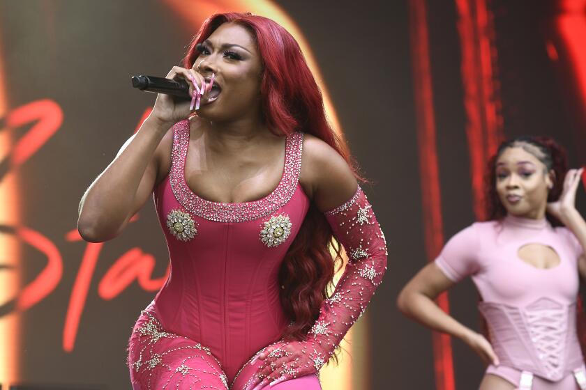 Megan Thee Stallion is rapping into a microphone on stage while dressed in a pink jumpsuit with silver patterns