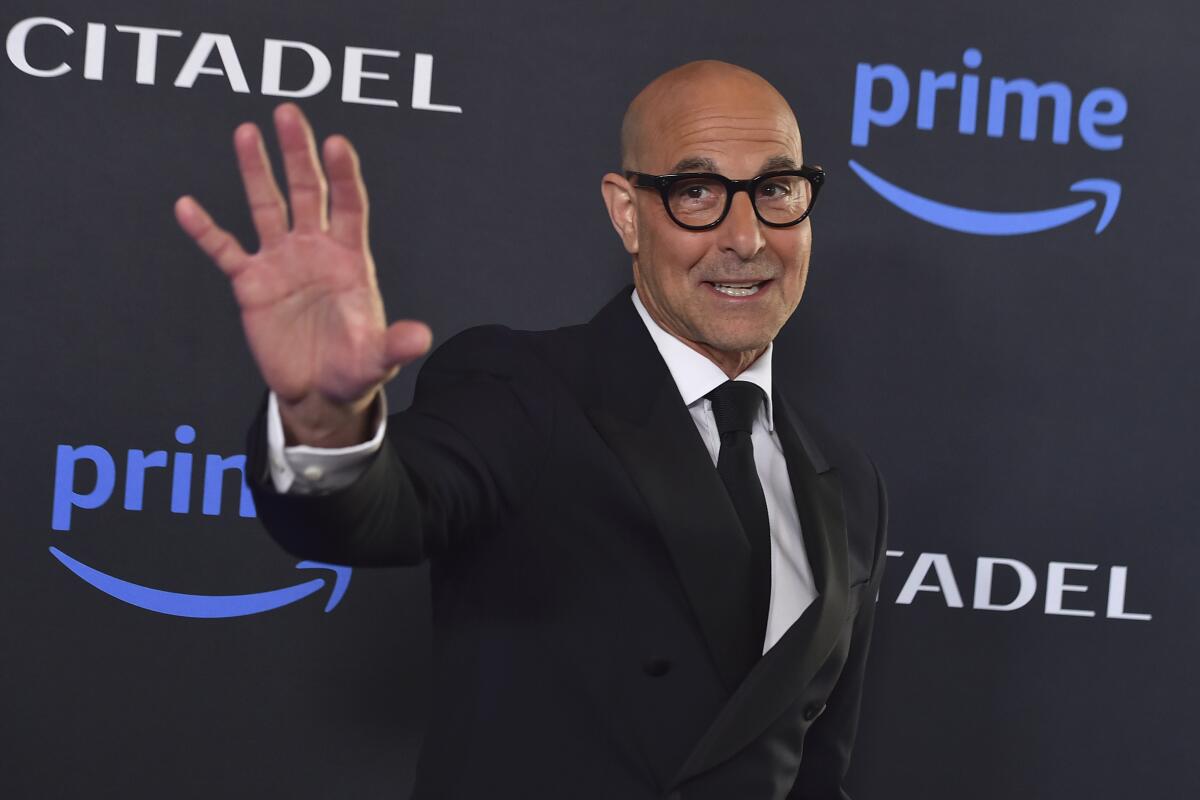 Stanley Tucci in thick black glasses and a black suit waving his right hand
