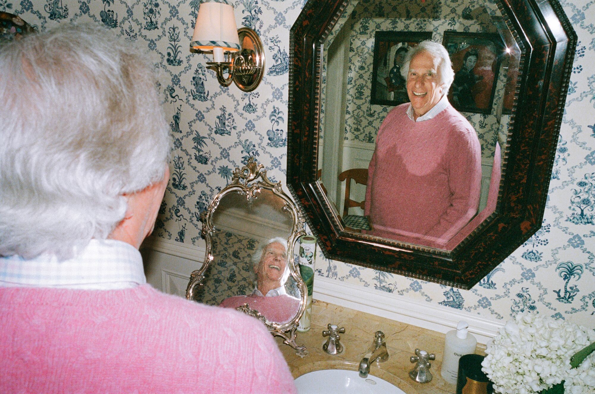 A man in a pink sweater looks at the camera in mirror