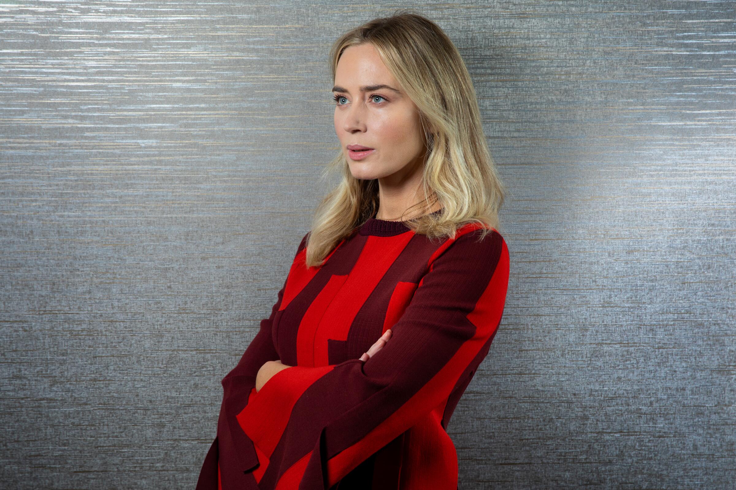 Emily Blunt stands with arms crossed, wearing a red and plum colorblock dress.