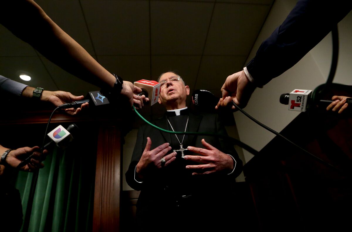 Seen from a low-angle view, a man wearing a black priest robe and a silver Christian-cross necklace speaks to reporters.