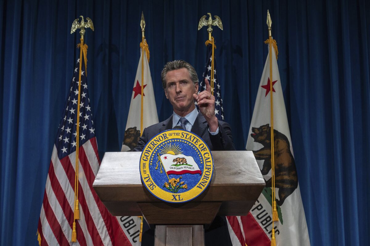 Gavin Newsom points a finger upward while speaking at a lectern in front of U.S. and California flags