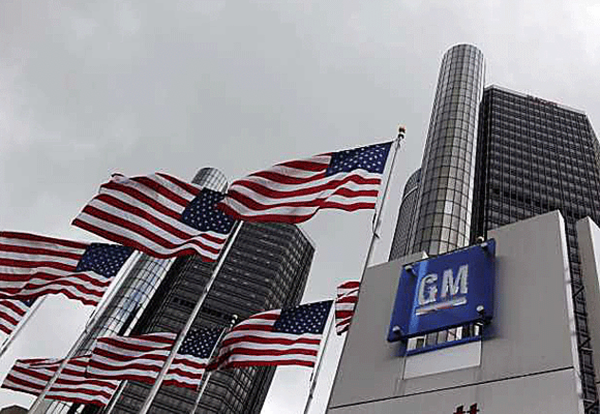A General Motors logo in front of two skyscrapers with American flags flying next to it.