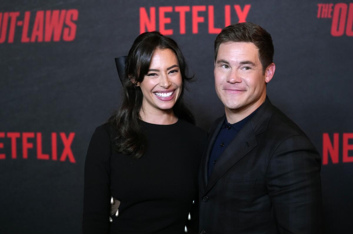 Adam Devine wears an all-black suit and Chloe Bridges wears a black dress as they pose for photos at a red carpet event