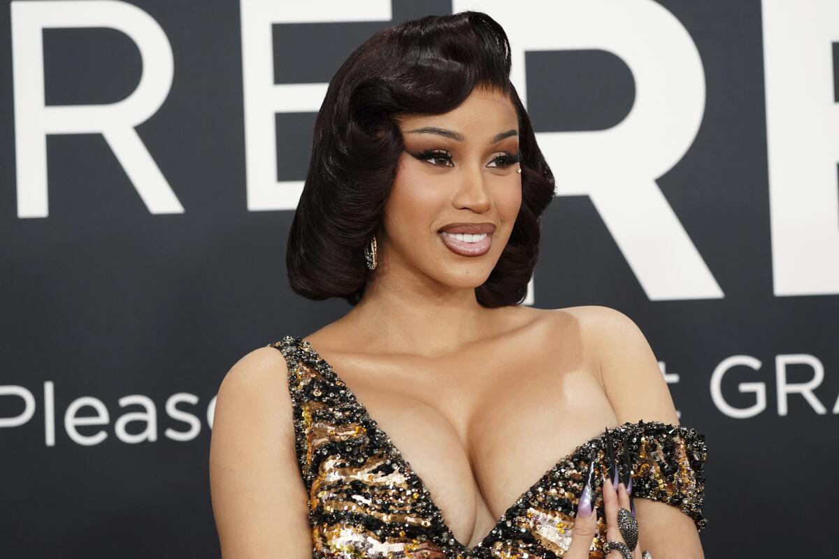 Cardi B in a golden gown smiling and posing against a black backdrop with white text