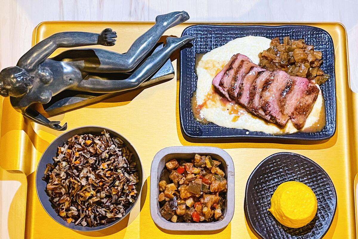 A gold tray holds dishes including sliced steak, with a statuette of a nude woman