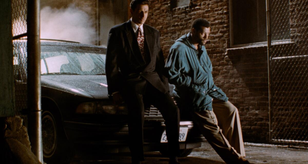 Two men lean on the hood of a car in an alley.