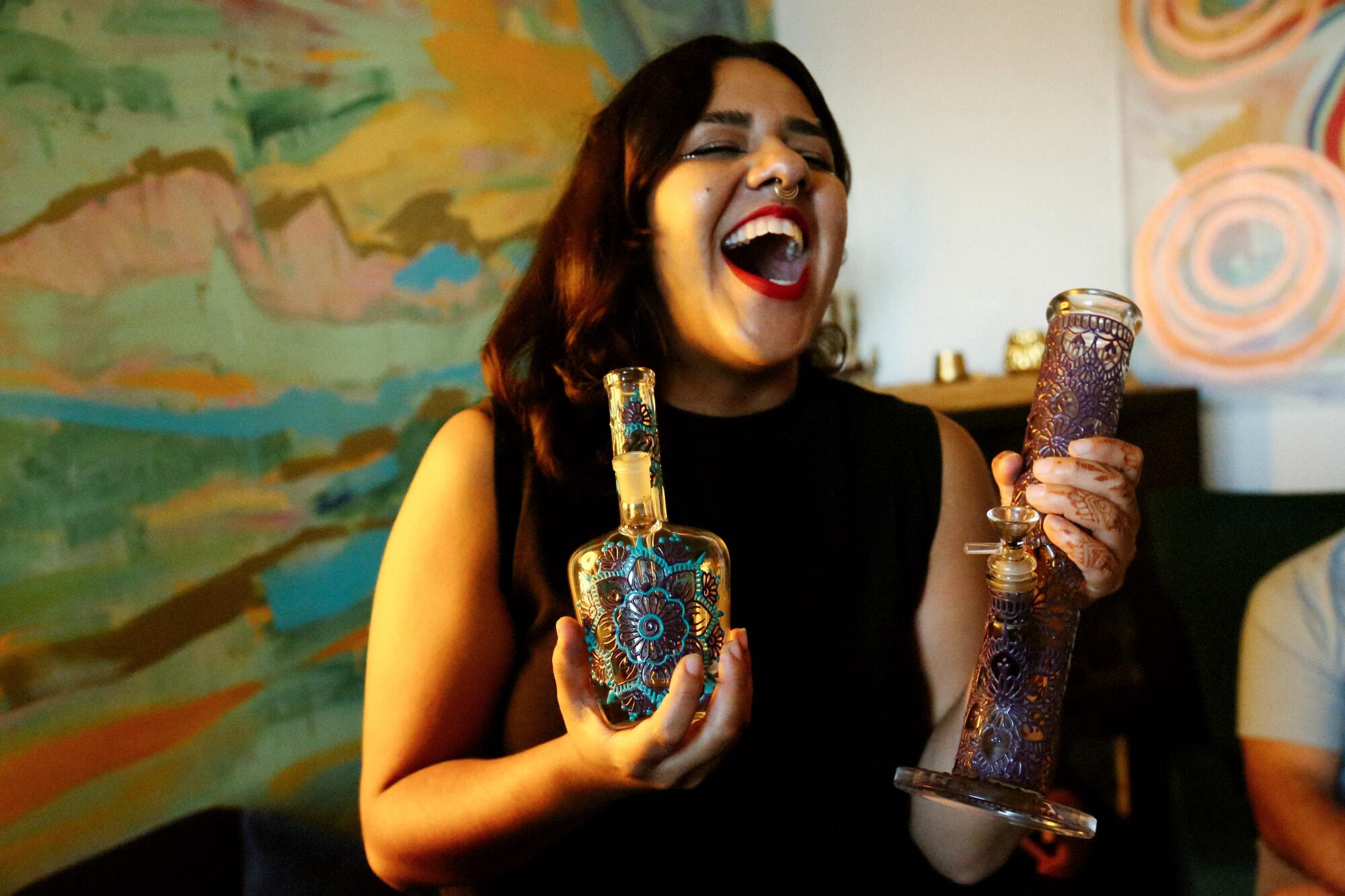 A woman laughing and holding a highly decorated glass pipe in each hand.