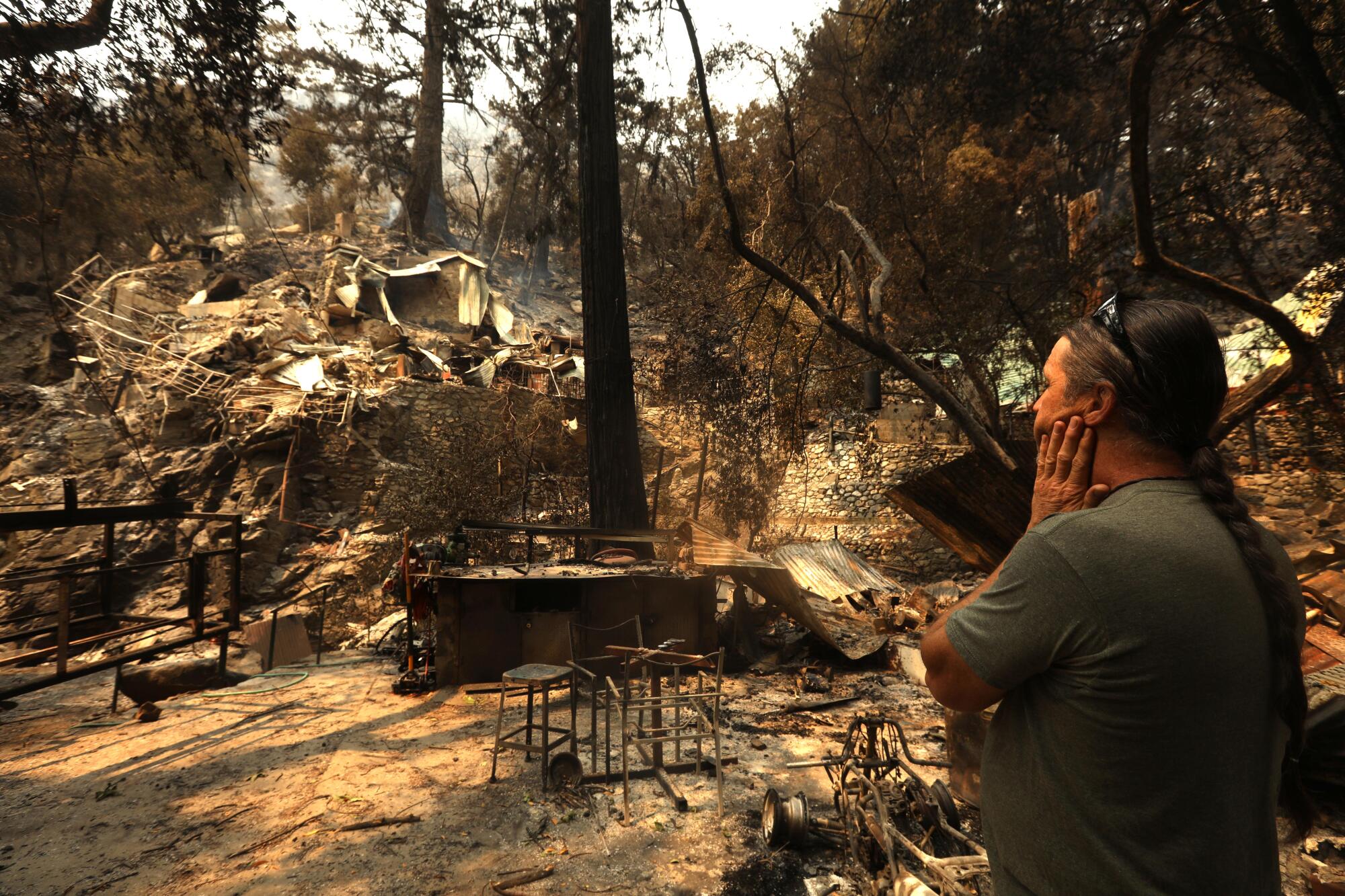 A man standing near scorched trees, furniture and rubble, holding a hand to his face.