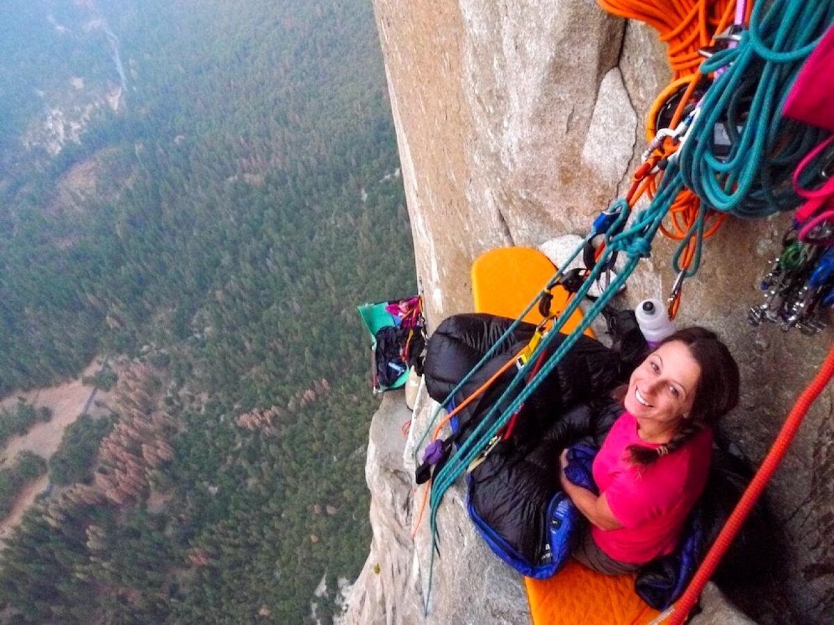 A smiling woman hangs by lines from the side of a mountain.