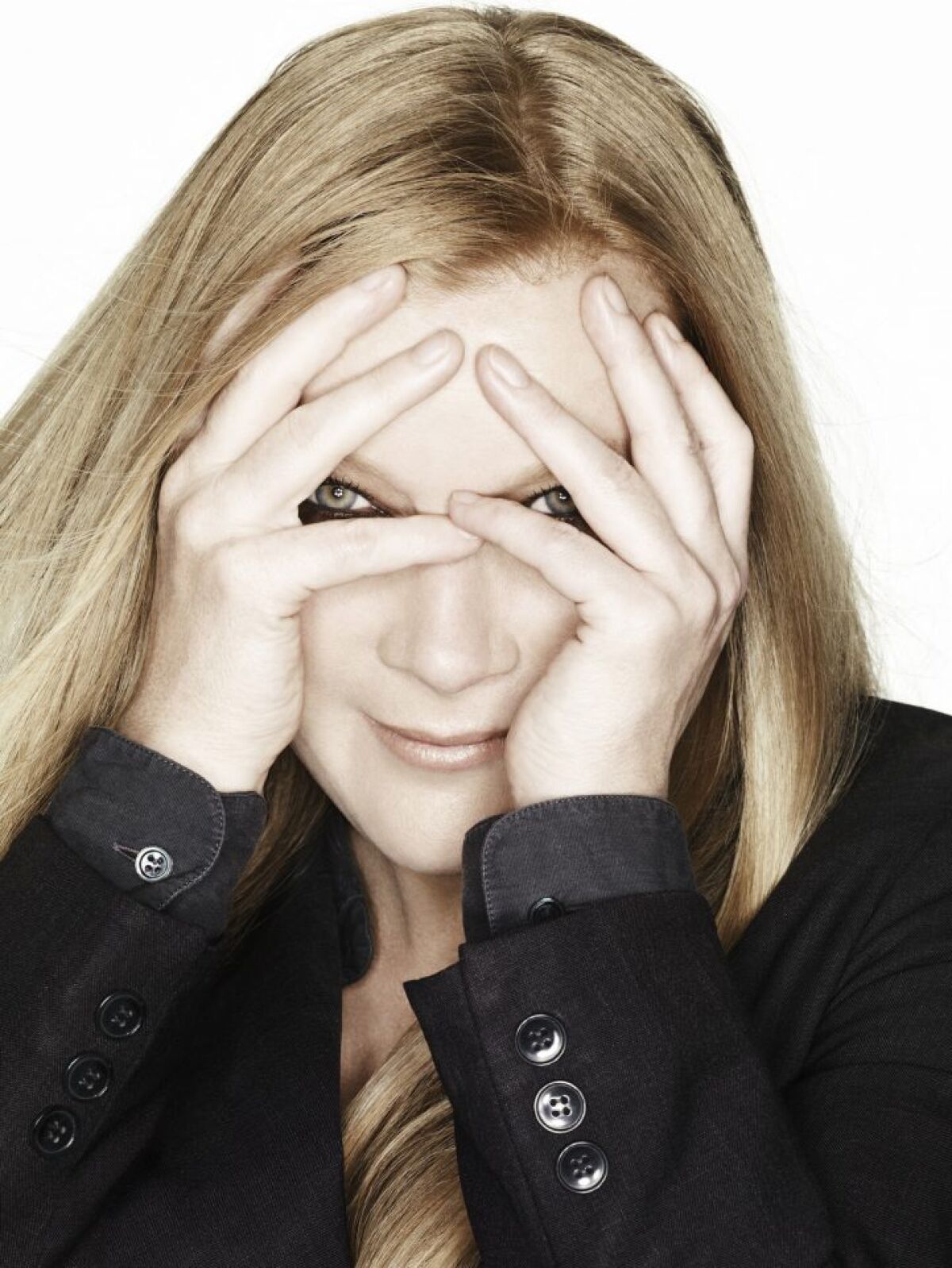 A smiling blond woman partially covers her face with her hands and peeks through her fingers.