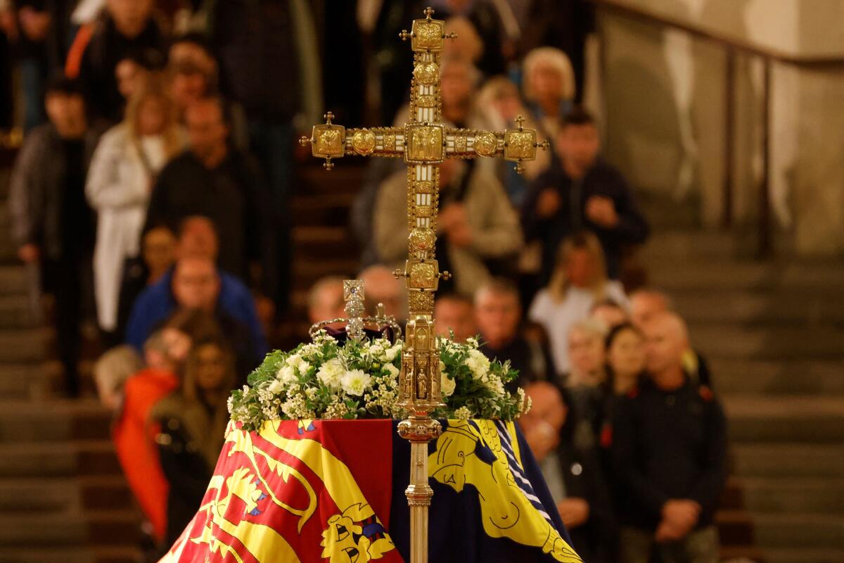 Mourners look at a coffin draped in a flag, with flowers and a crown on top of it, accompanied by a golden cross.