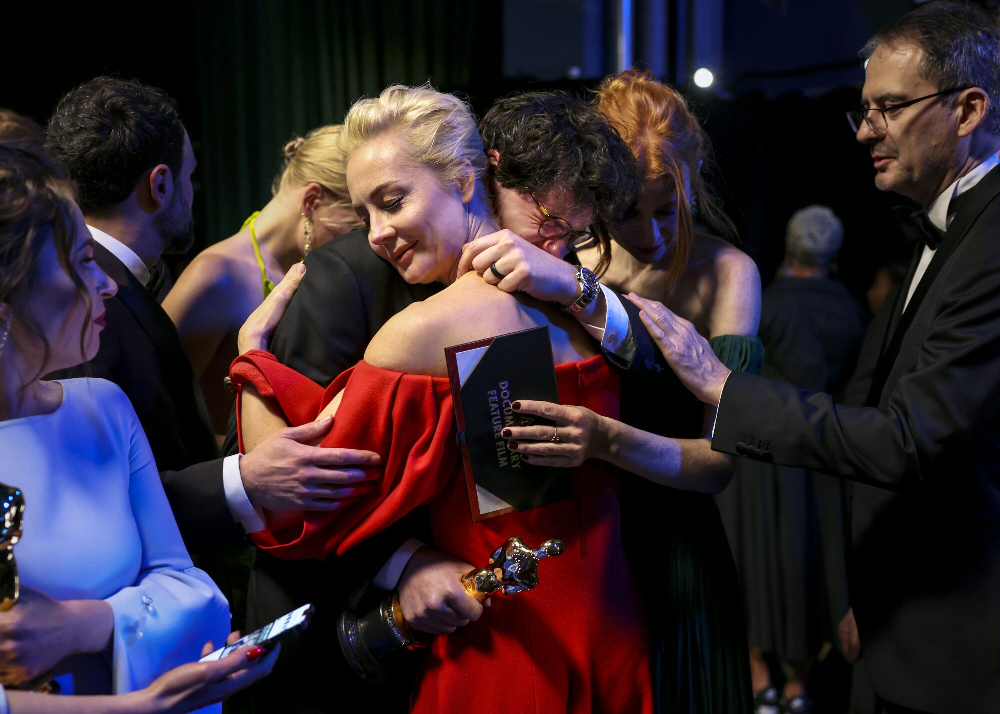 A group of people in evening wear hug while one holds an Oscar statue. 