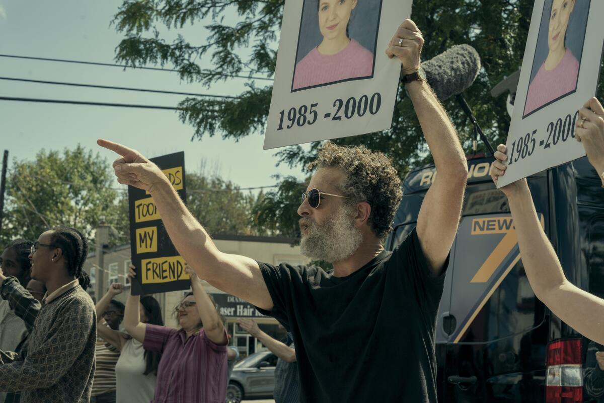 A man in black T-shirt points his finger upward and holds a sign with the photo of a girl and the years 1985-2000.