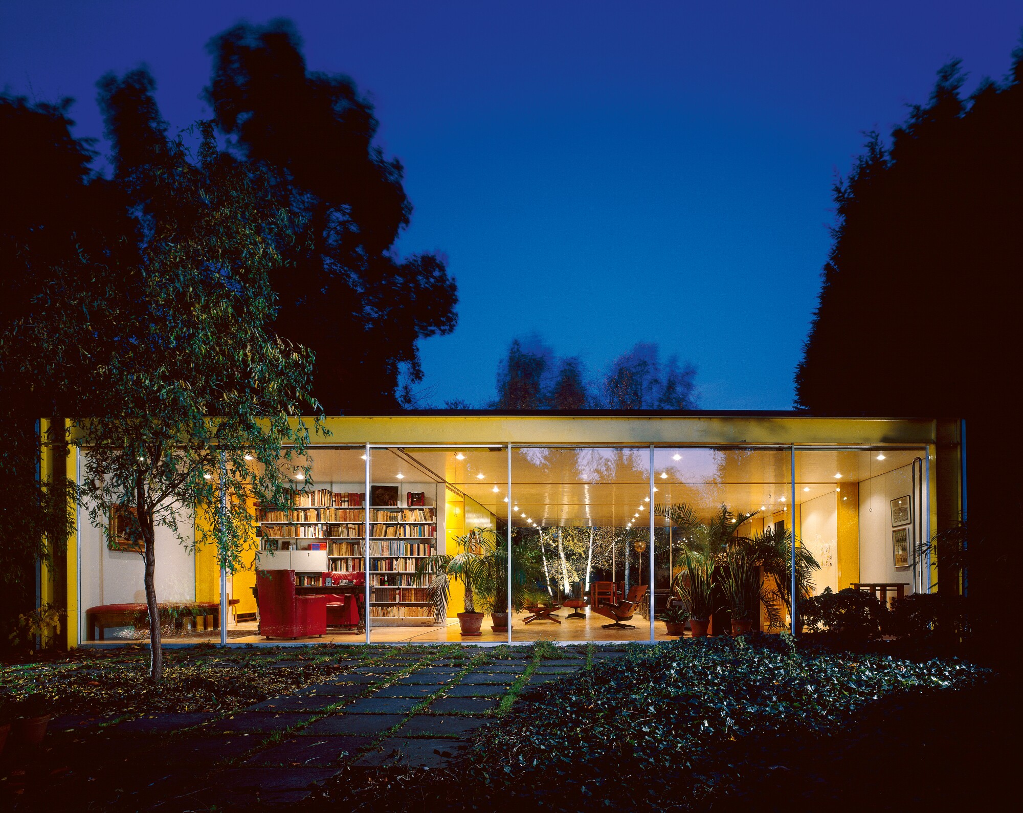 A glass facade of the Rogers house reveals the living area at dusk — all framed by the home's bright yellow steel beams.