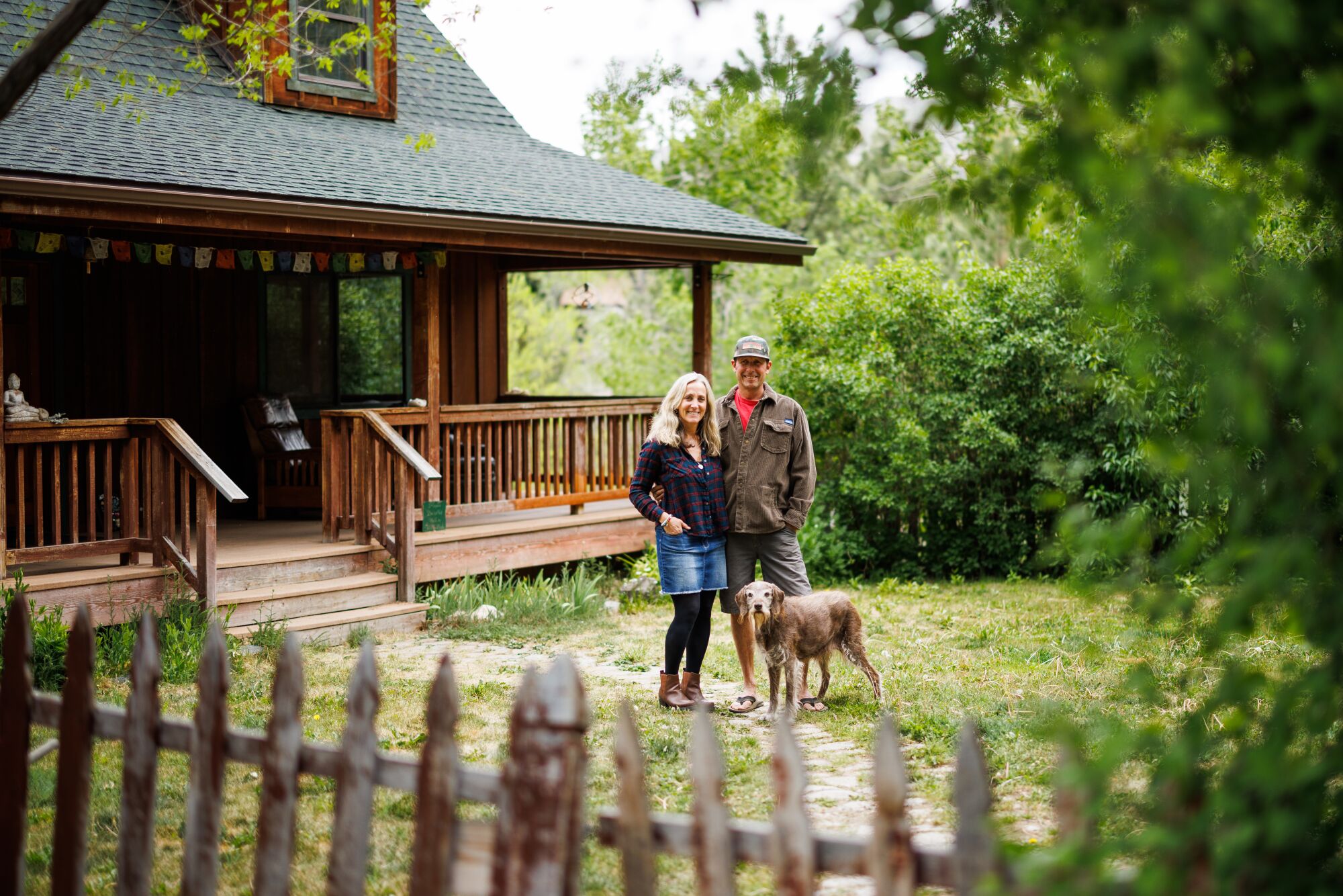 A woman, man and dog standing outside a wood home surrounded by trees
