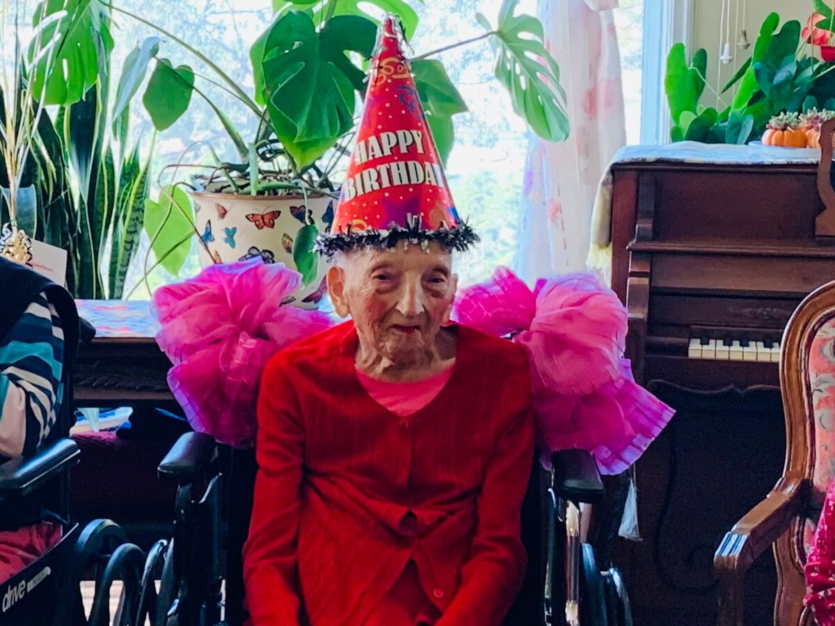 An elderly woman sits in a wheelchair while wearing a large Happy Birthday cone hat