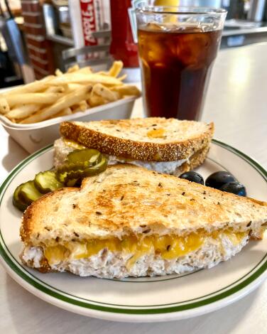 A tuna melt sandwich cut in half with pickle chips, black olives, fries and a soda