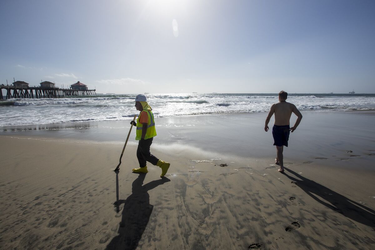 A visitor to Huntington Beach from Salt Lake City enters the water as waste management employee combs the beach for oil.