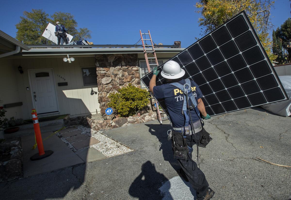 A worker in a hard hat carries a large solar panel toward a single-story house.