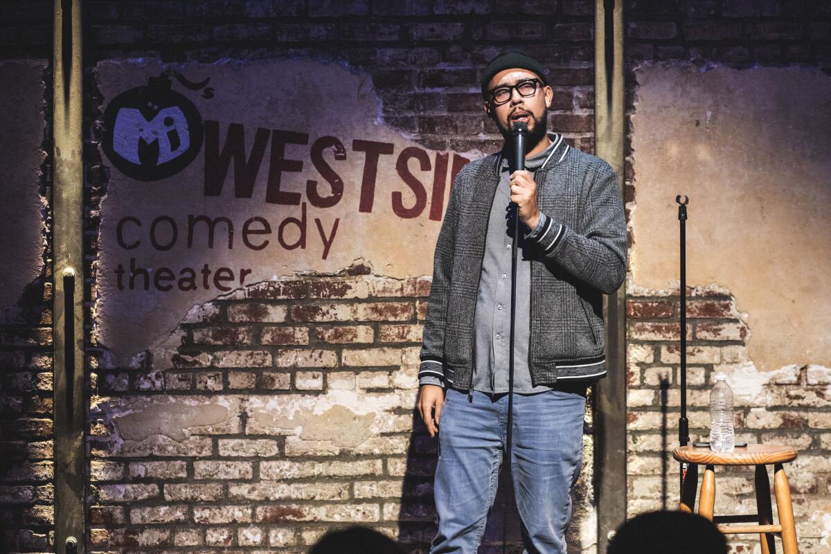 A man in jeans, gray T-shirt and gray jacket with a handheld mic onstage at Westside Comedy Theater