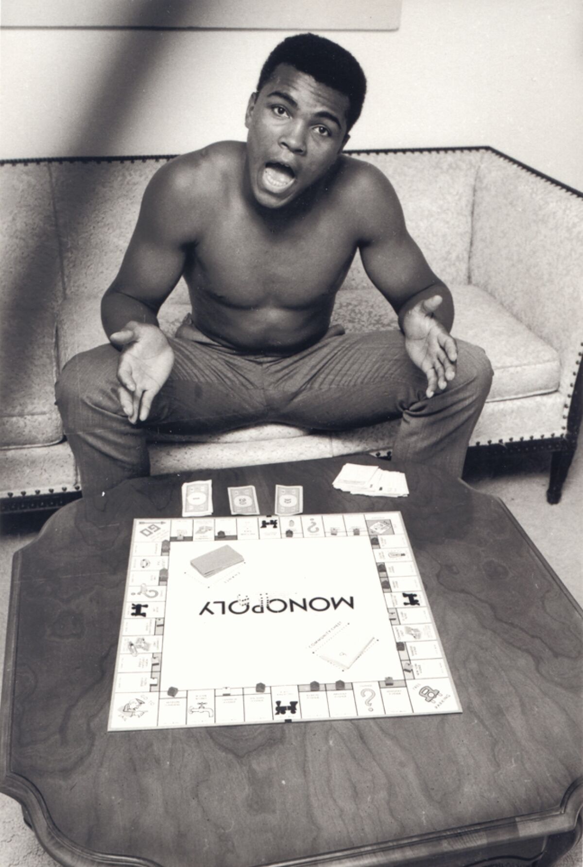 A shirtless man plays the board game Monopoly.