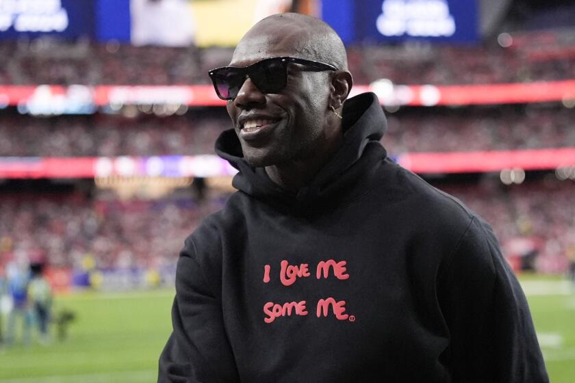 Terrell Owens smiles while wearing shades and a hoodie that reads "I Love Me Some Me" and standing on the field before a game