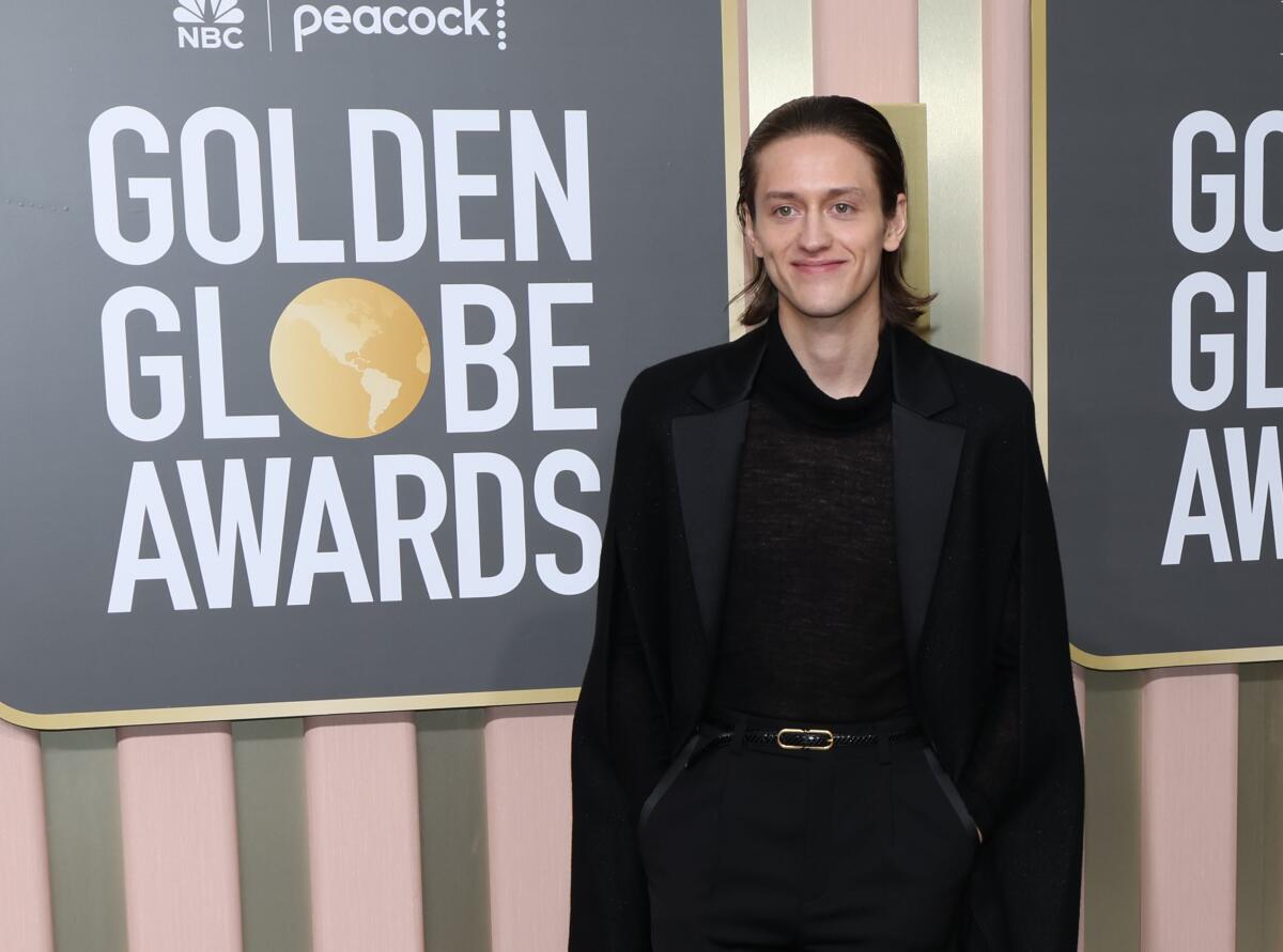 Percy Hynes White is smiling on the Golden Globes red carpet and is wearing an all-black suit with a long coat