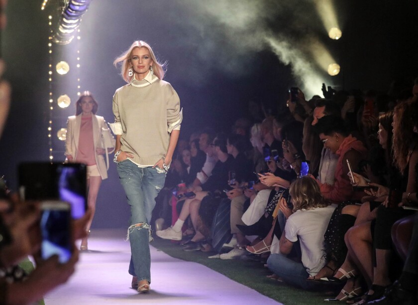 A woman walks the runway in a look from Brandon Maxwell: beige sweater over a white collared shirt and blue jeans, ripped at the knee.