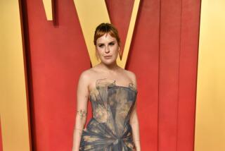 A woman with short red bangs posing in a strapless, blue and gold gown on a red carpet