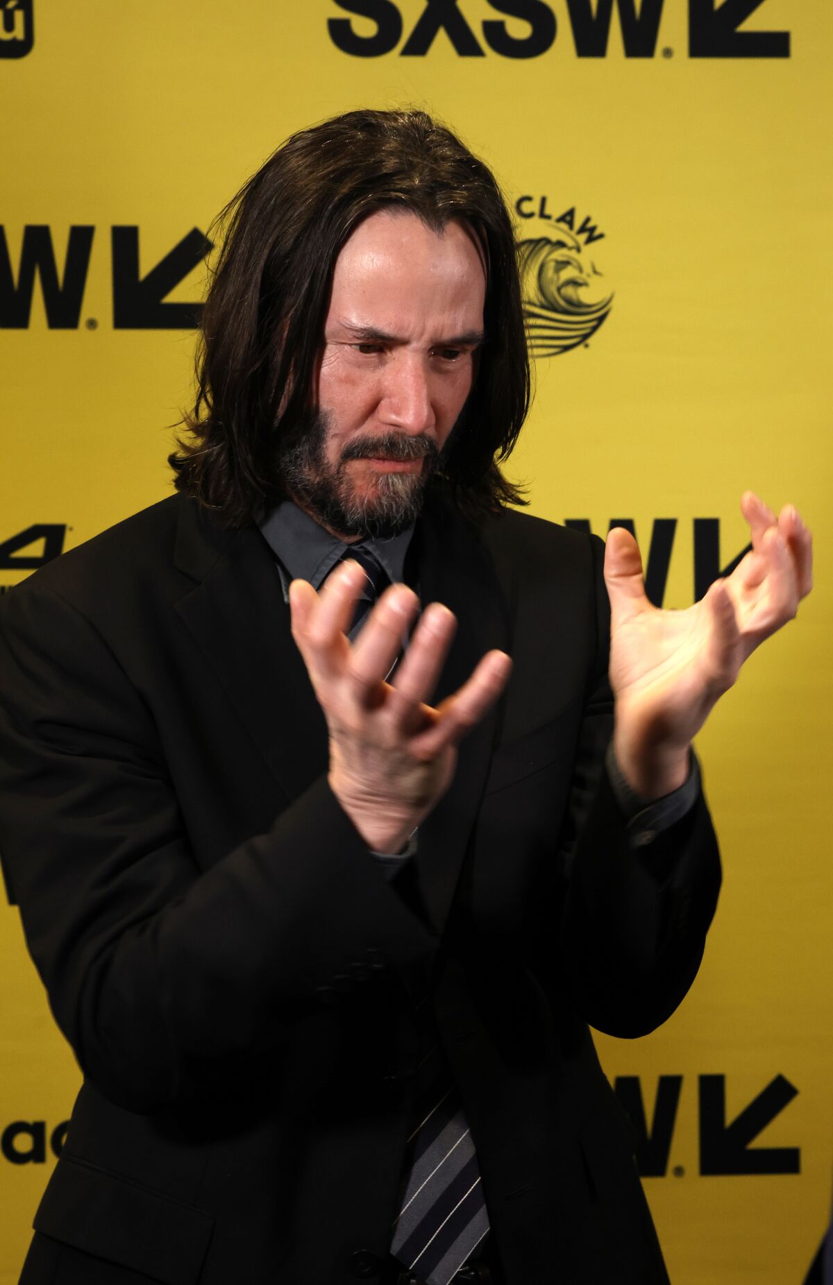Keanu Reeves in a dark suit and tie against a yellow background with various brand logos