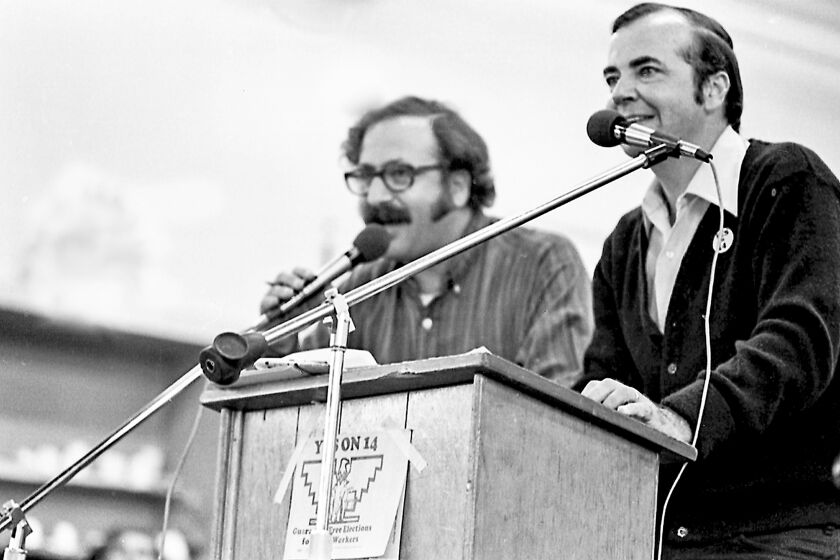 A black-and-while image of two men speak to microphones from a podium.