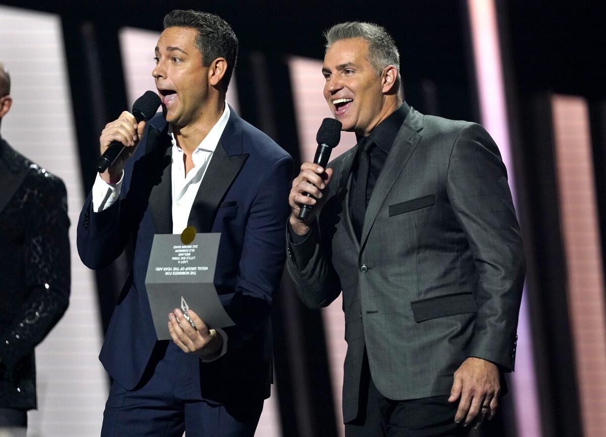Two men dressed in suits hold microphones, one of them with an open envelope in the other hand. 