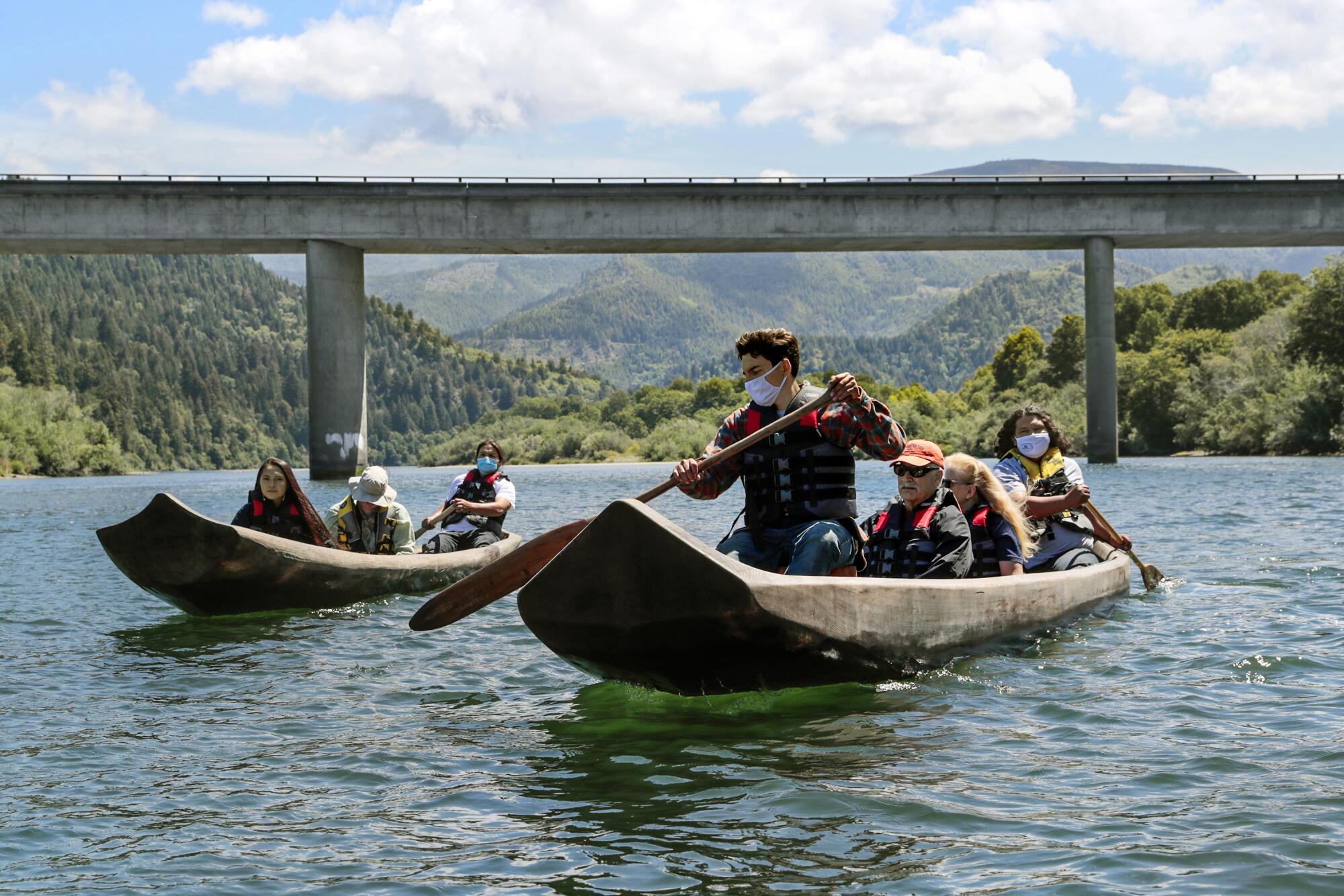 Boaters in dugout canoes pass near a bridge over a forested lake