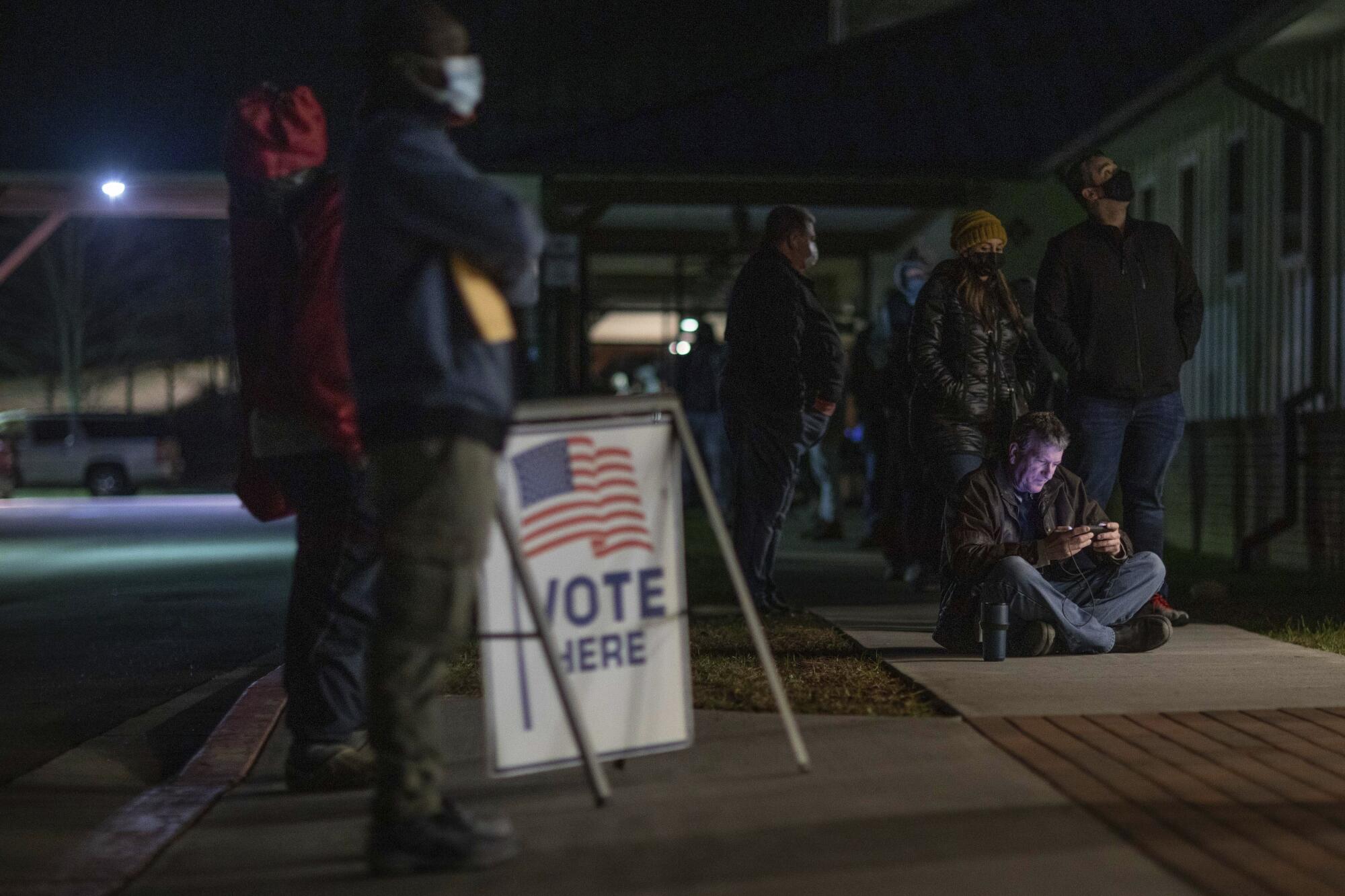 Voters wait in the dark by a "Vote here" sign, all in masks except one man sitting on the ground looking at his phone.