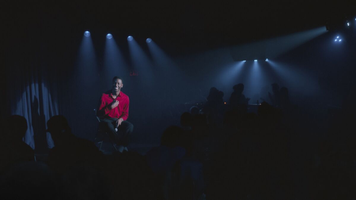 A man in a red shirt sits on a spot-lighted stage