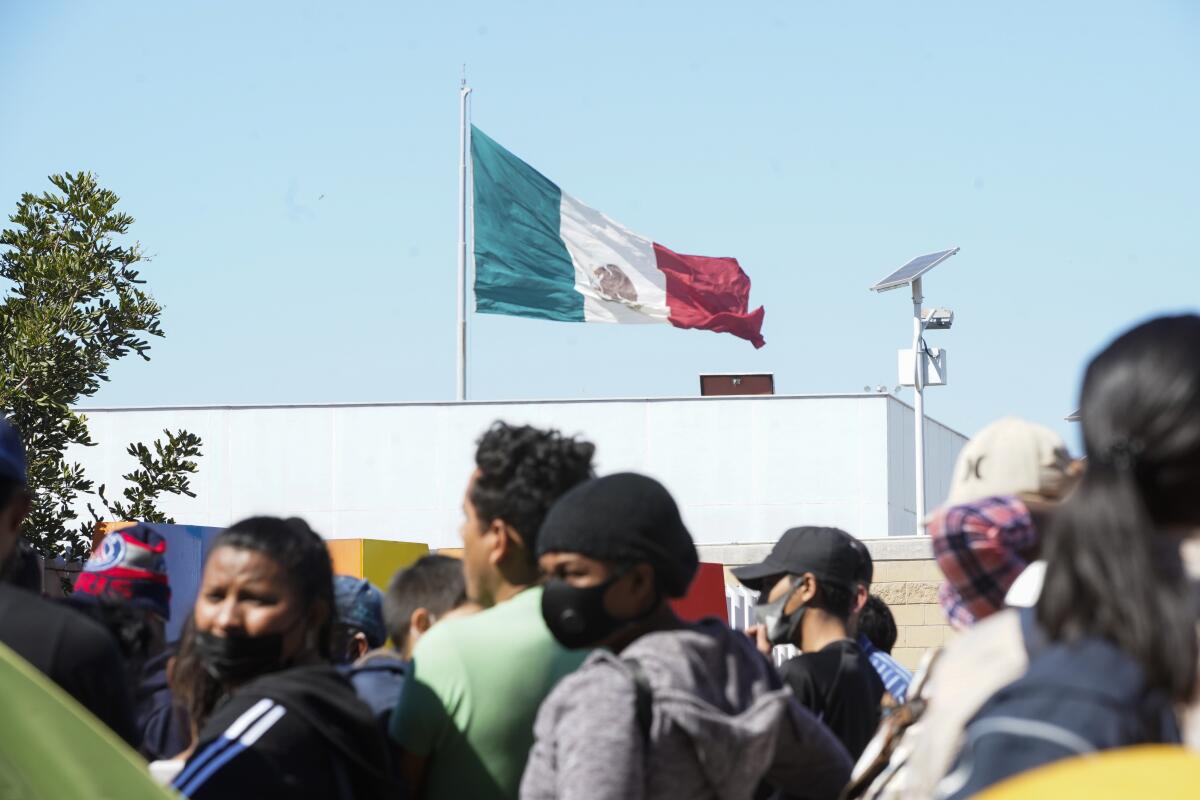 Asylum seekers stand in El Chaparral plaza below the Mexican flag