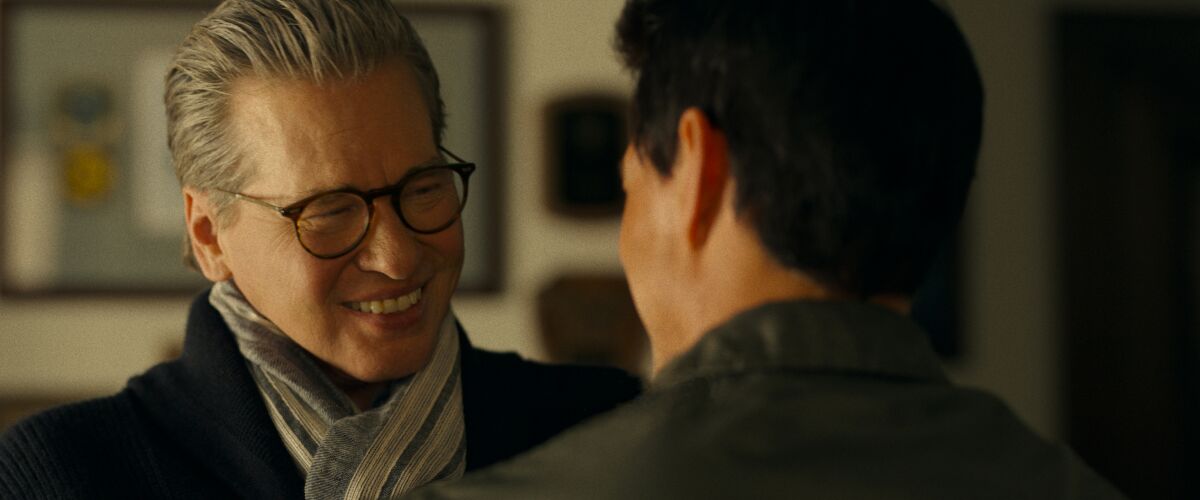 A man in glasses and a scarf smiles at a man facing him.