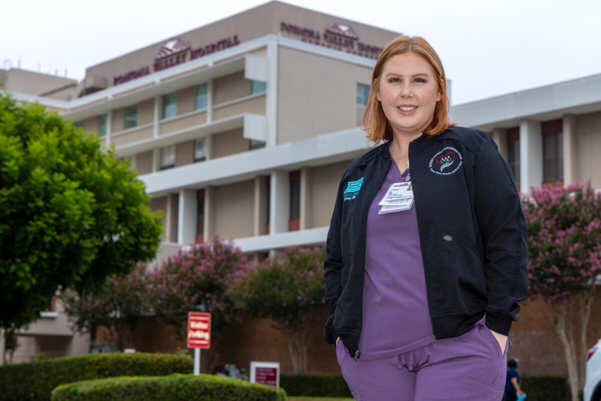 A woman stands wearing a purple top and pants and a dark jacket.