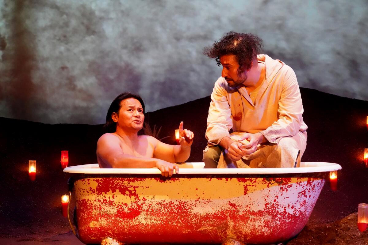A man sitting in a tub and pointing beside a monk perched on the edge of the tub