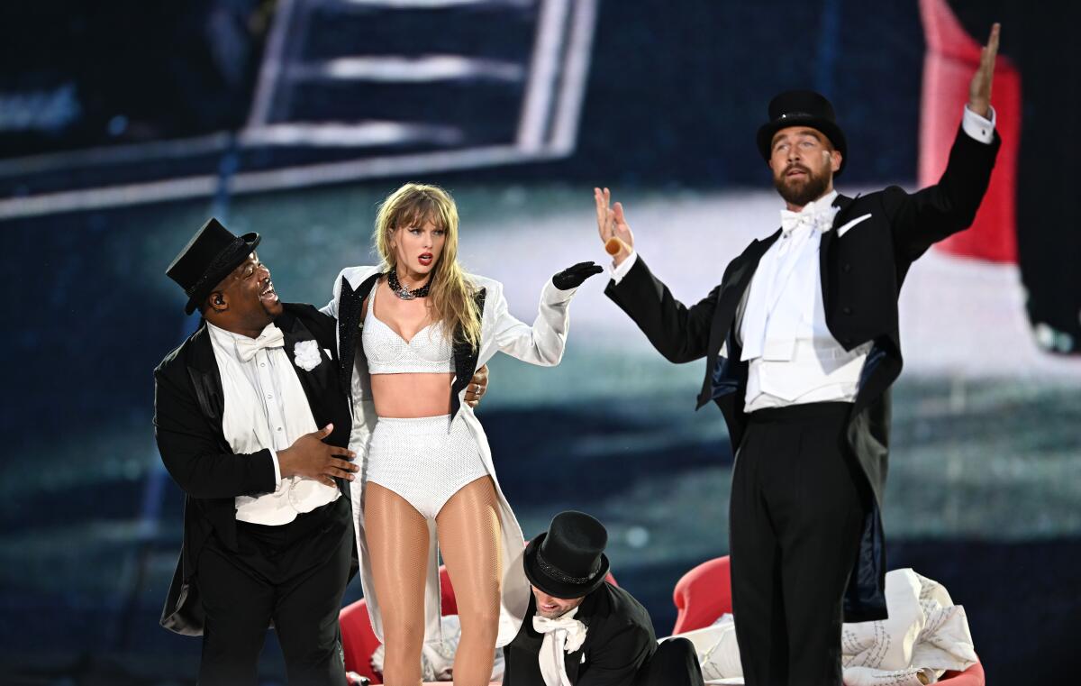 Taylor Swift performing onstage, with Travis Kelce in a top hat and tuxedo joining her