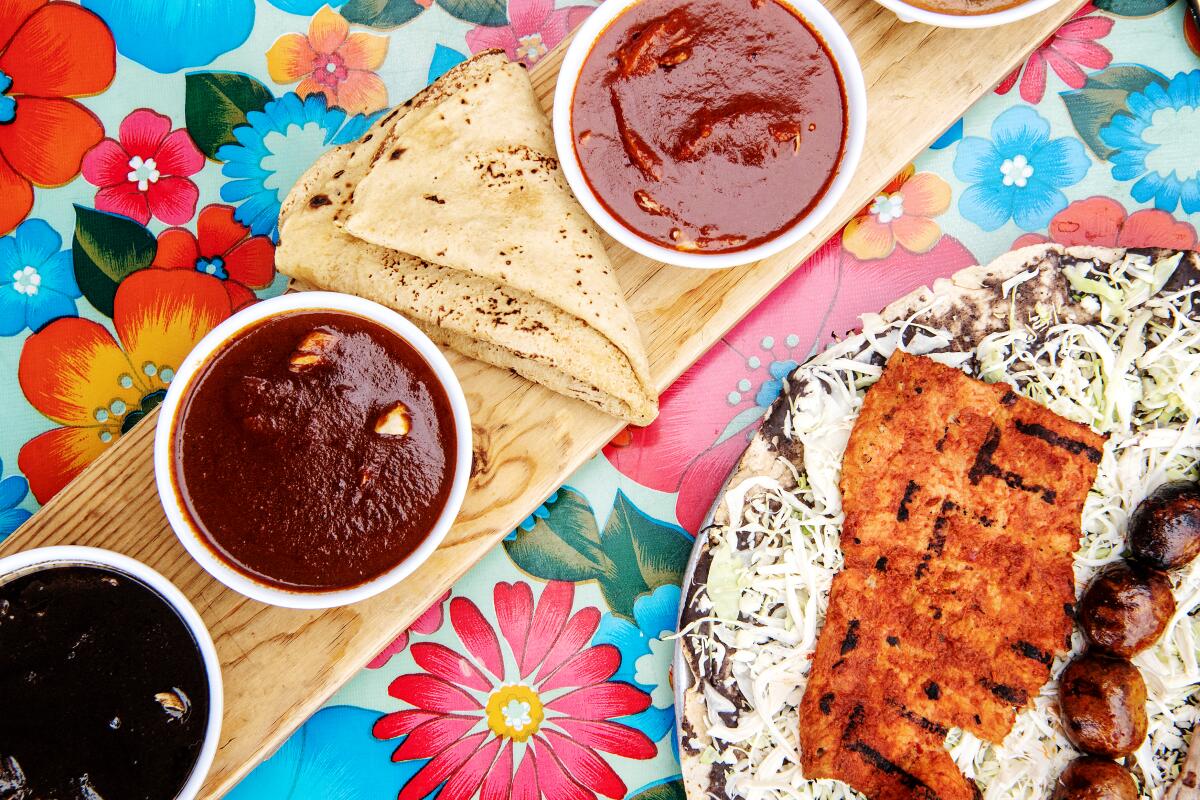Cups of different moles with tortillas next to a tlayuda Guelaguetza on a colorful floral tablecloth.