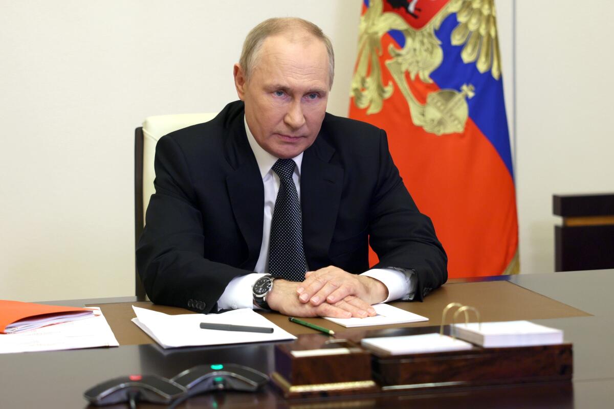 A grim-faced man in dark suit and tie, one hand over the other, seated at a desk with a flag behind him