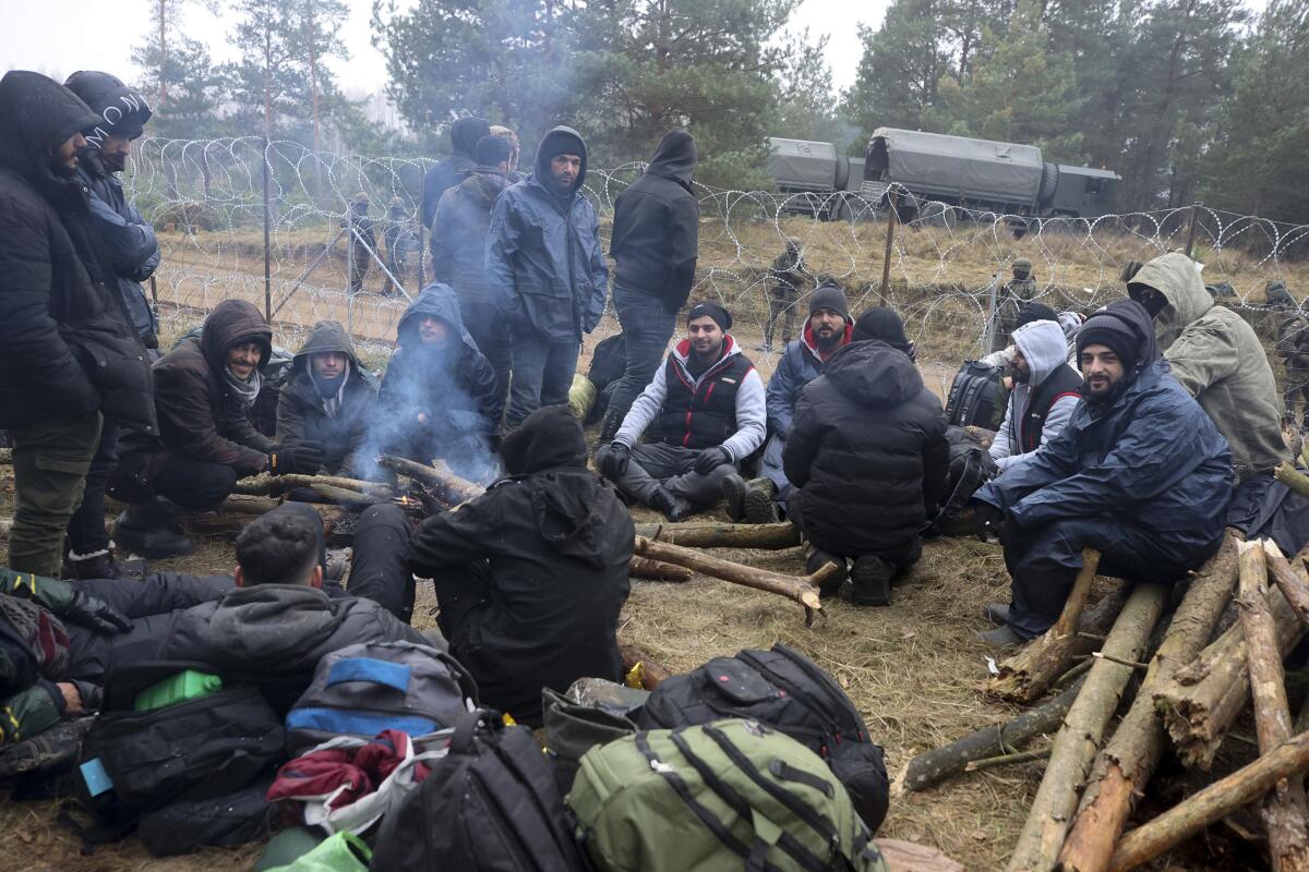 Men in winter coats and hats sit around a fire inside a fenced-off area with soldiers on the other side.