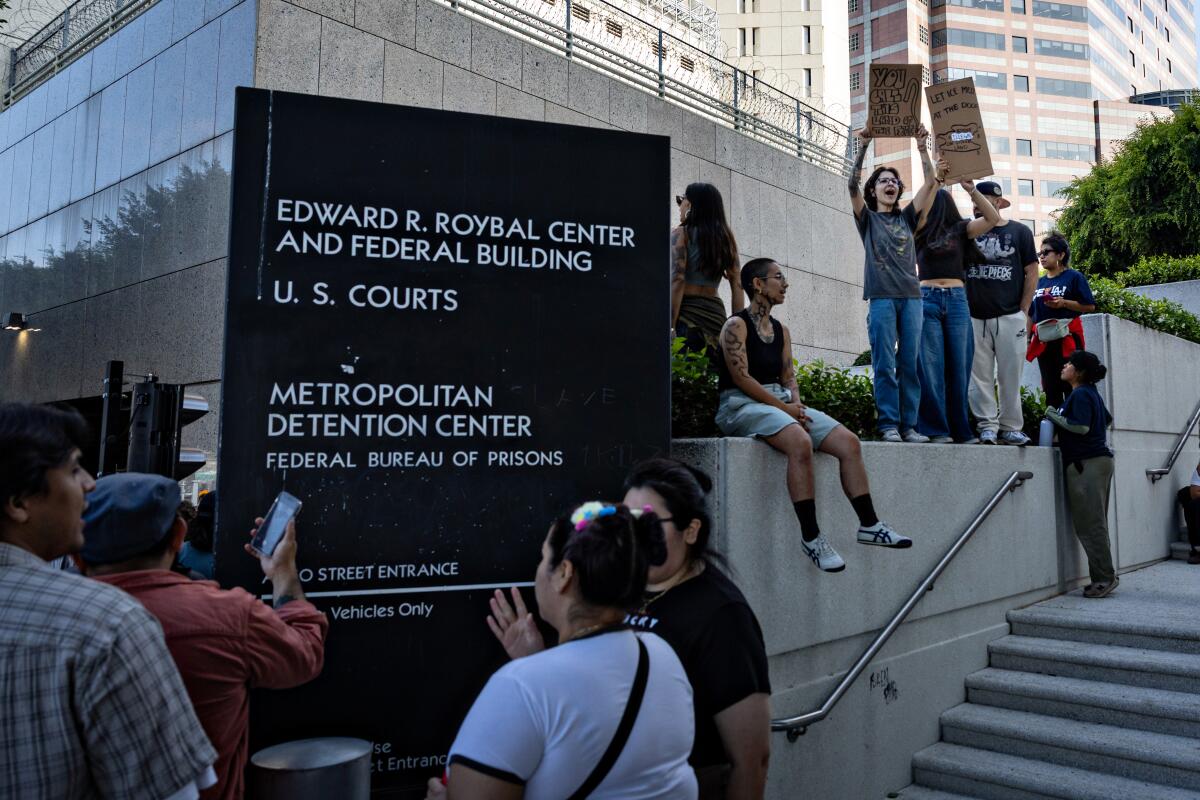 Demonstrators stand around and sit on a ledge outside the Edward R. Roybal Federal Building