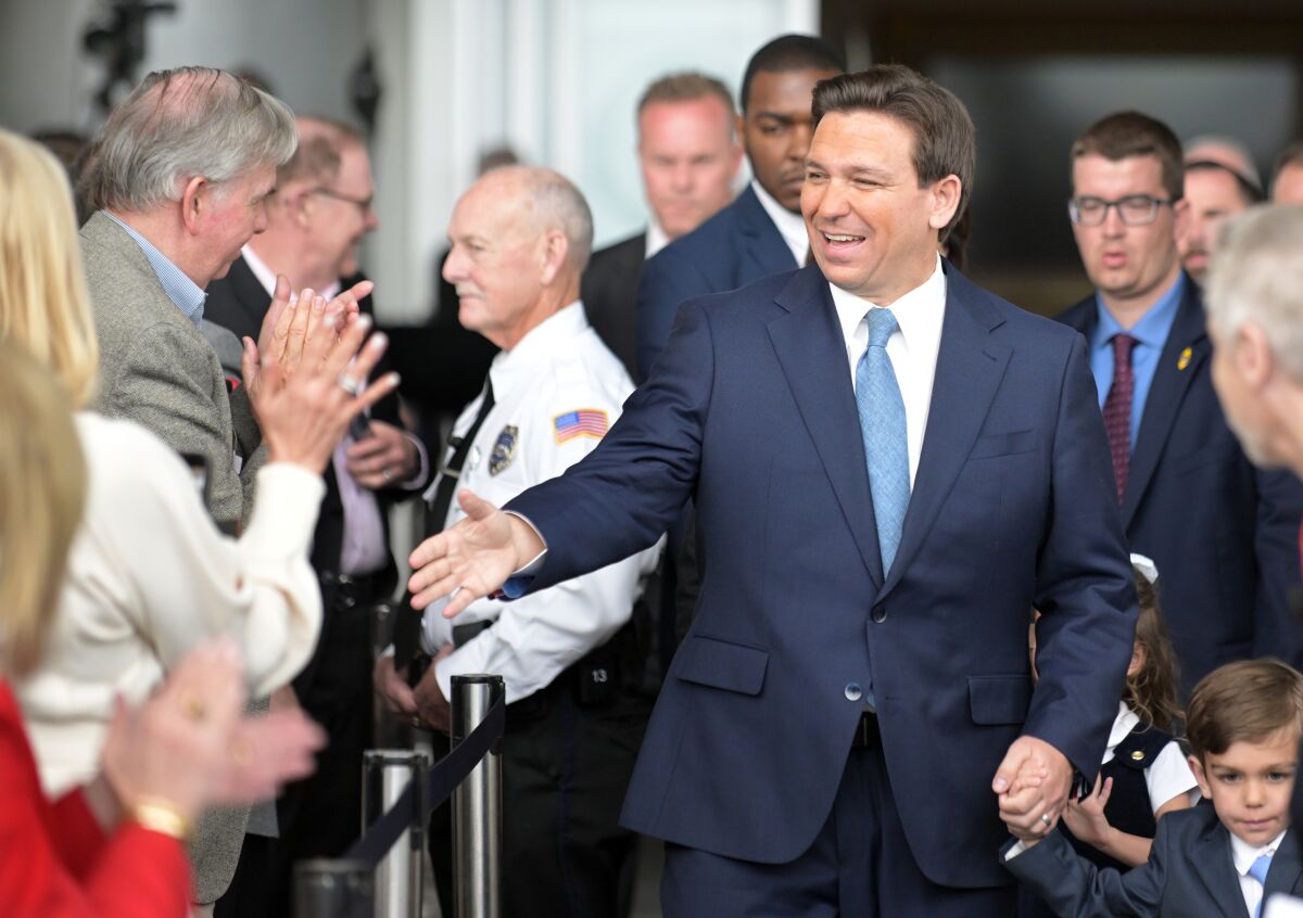 A man with dark hair, in a dark suit and blue tie, extends his hand toward people applauding