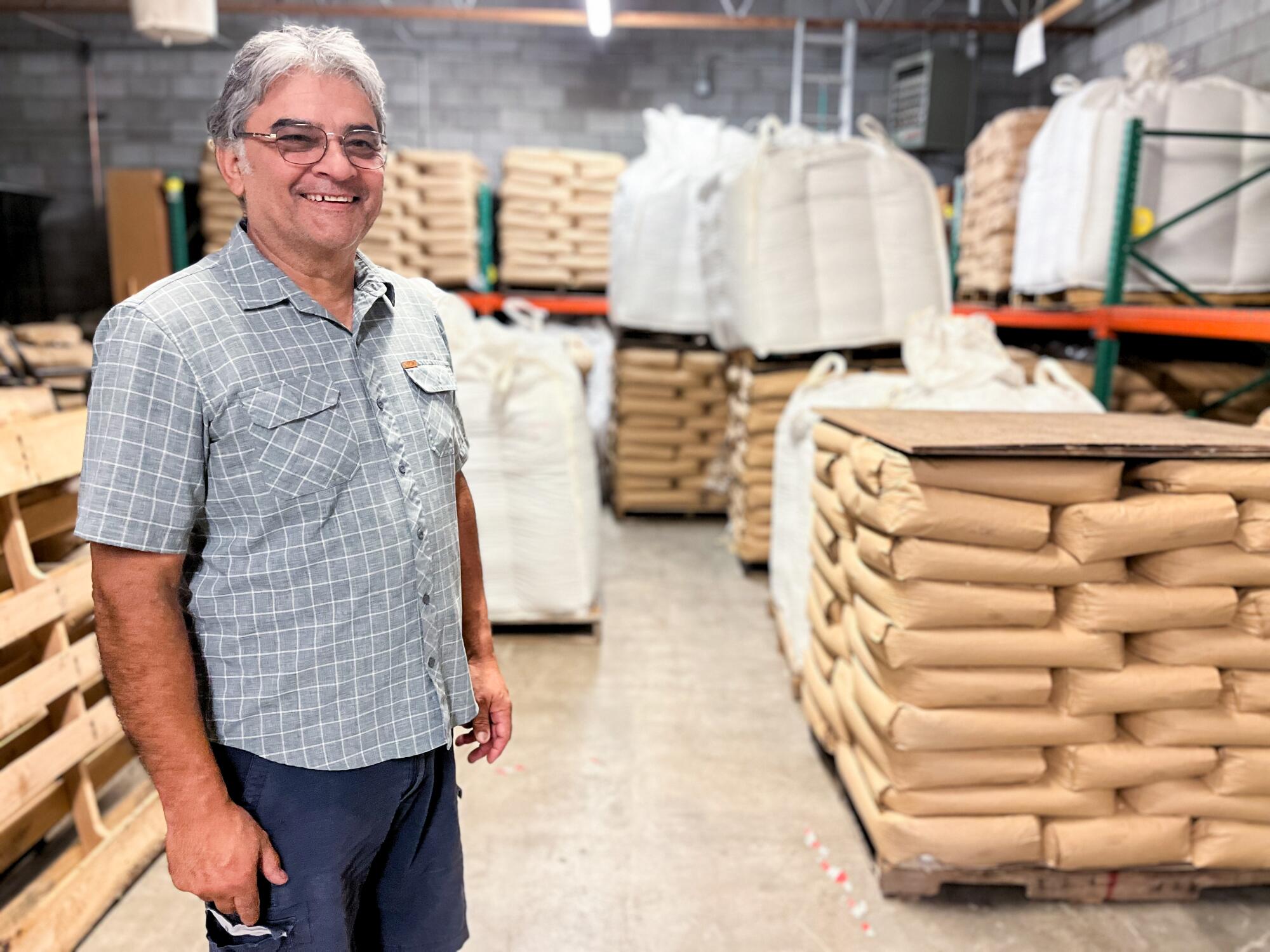 A man with gray hair, wearing glasses and a gray-blue checked shirt, smiles as he stands in a warehouse with bags on pallets 