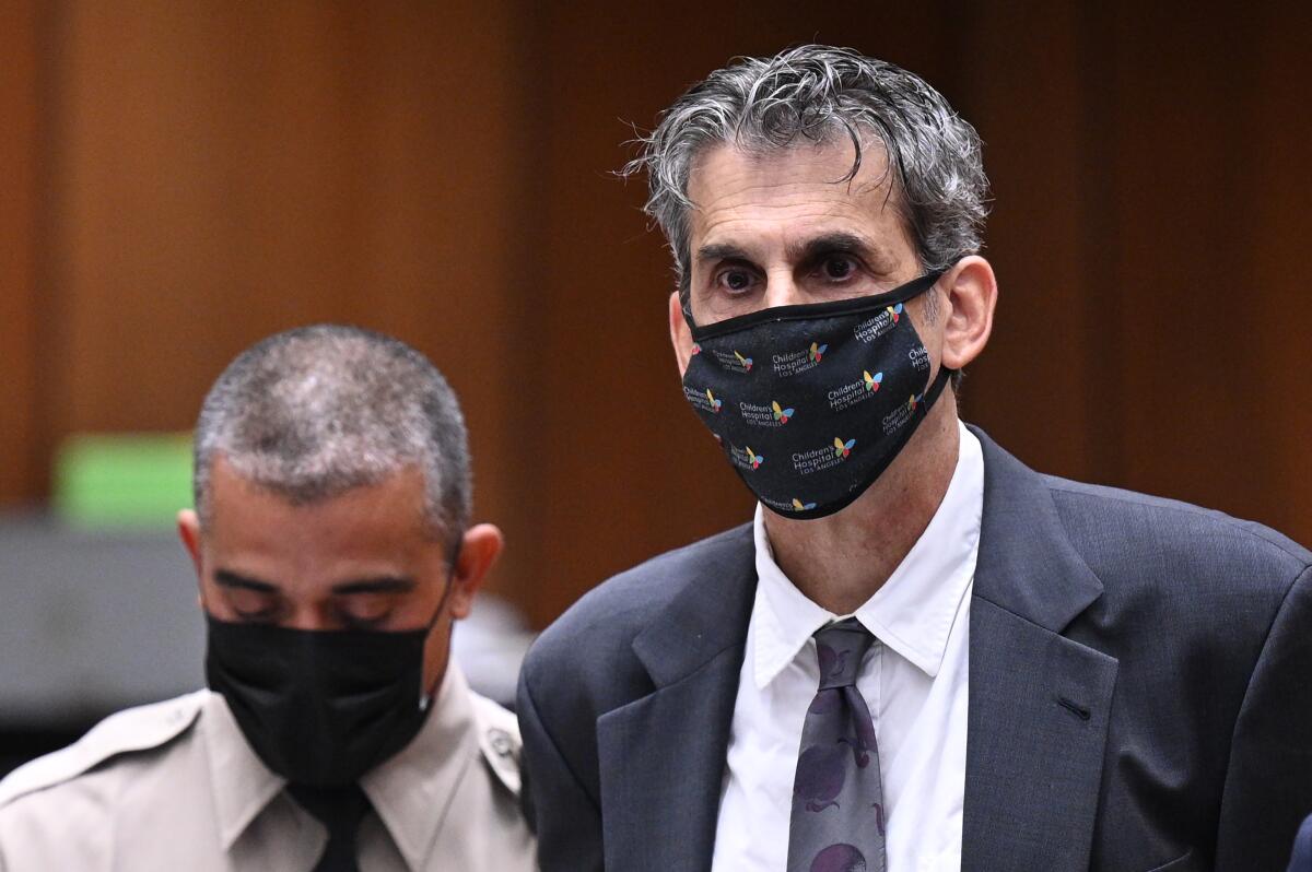A man with gray hair wearing a gray suit and a black patterned face mask