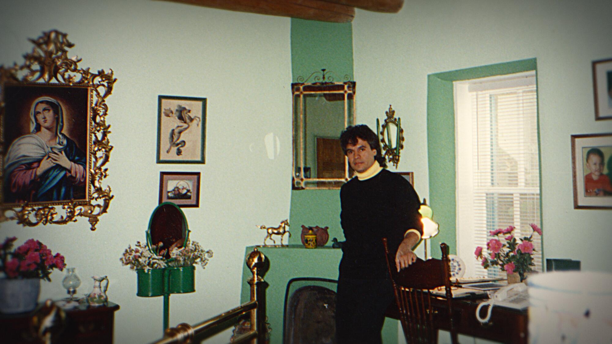 Man in a black sweater with a yellow button-up shirt and black pants stands in a light green room surrounded by wall decor